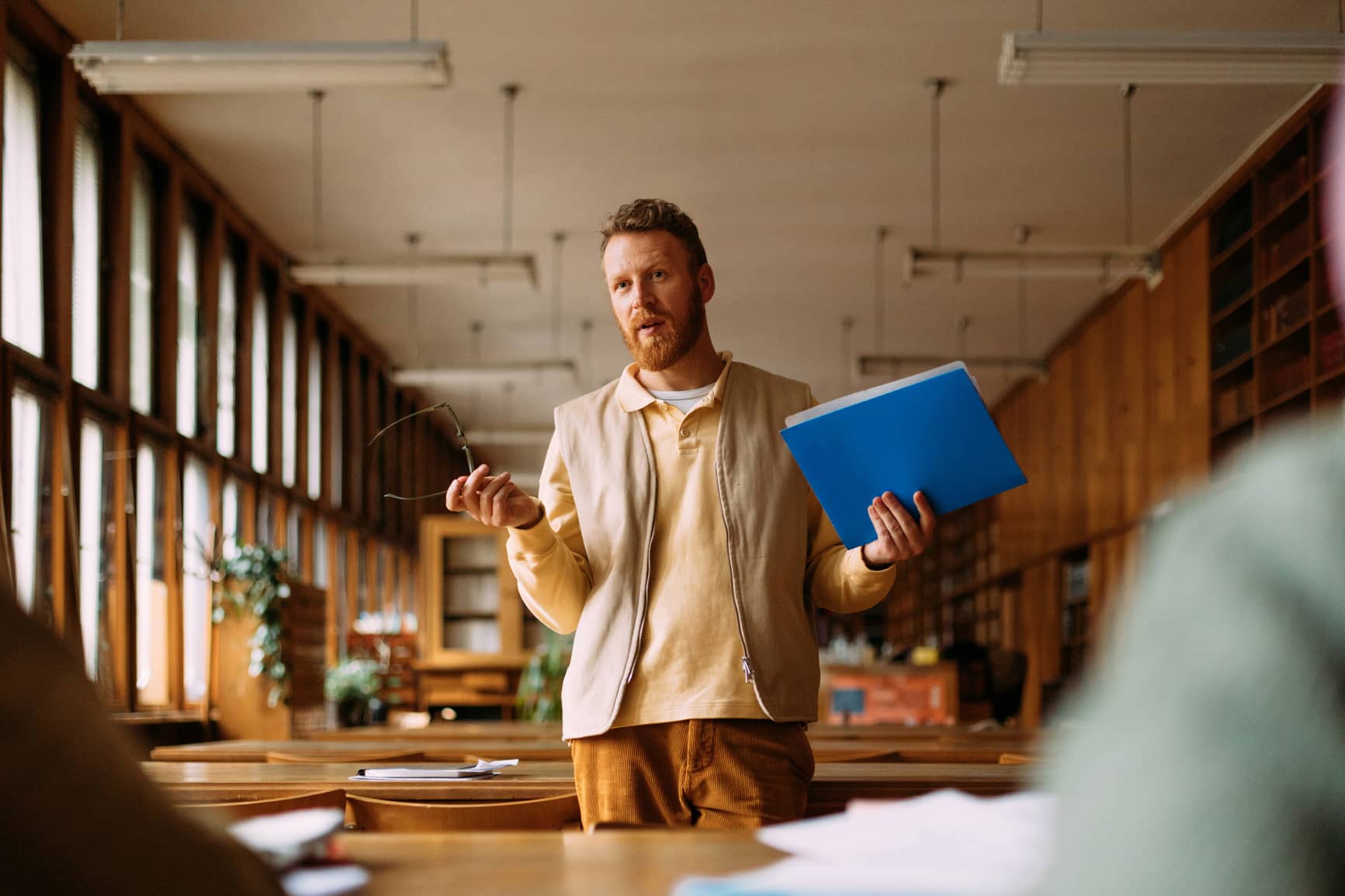 man standing in a library holding folder in his left and and glasses on his right hand